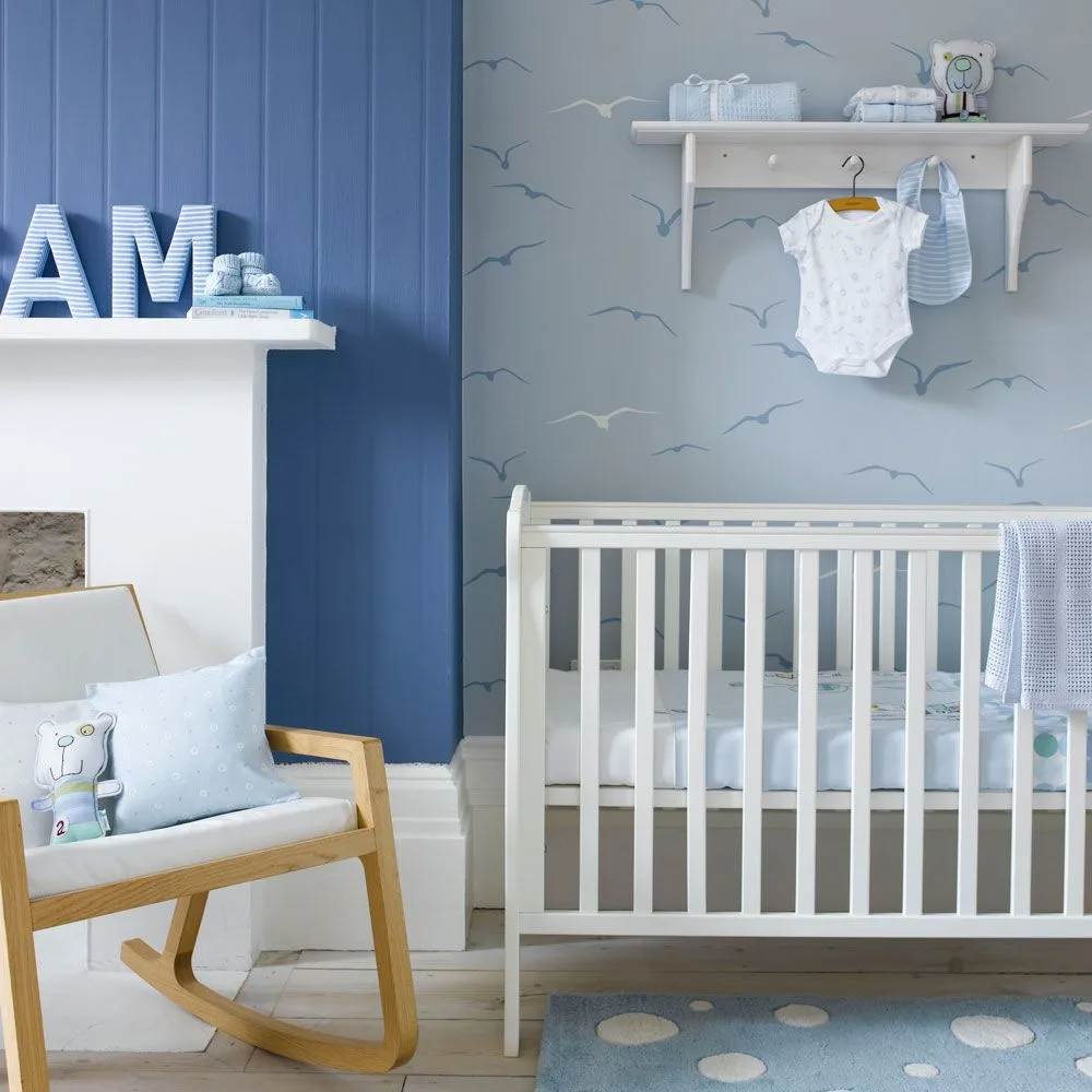 White crib against pale bird wallpaper beside deeper blue paneling, pale fireplace and wooden rocking chair with blue fabric.