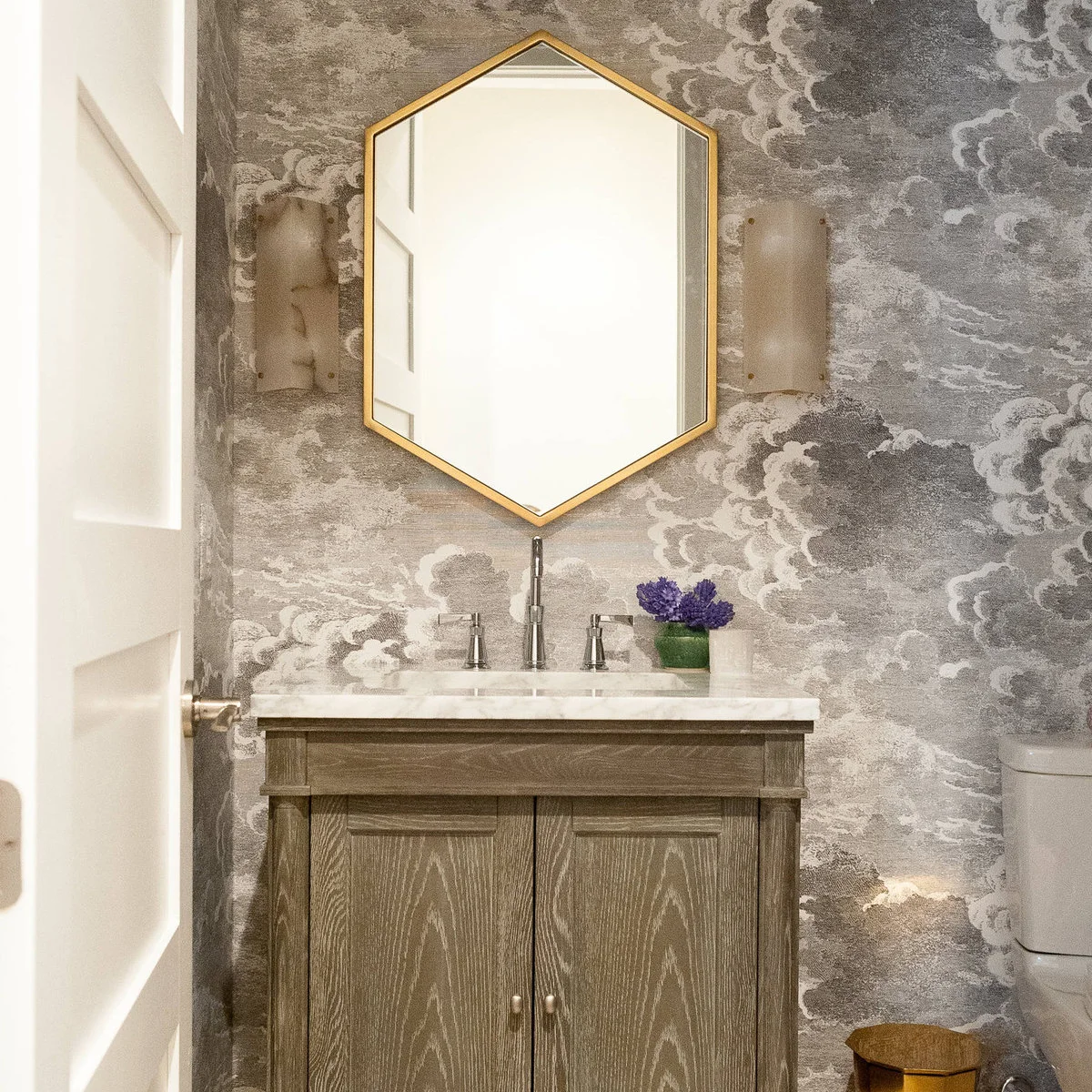 A wood vanity with a pale counter beneath a hexagonal mirror against cloud-patterned walls.