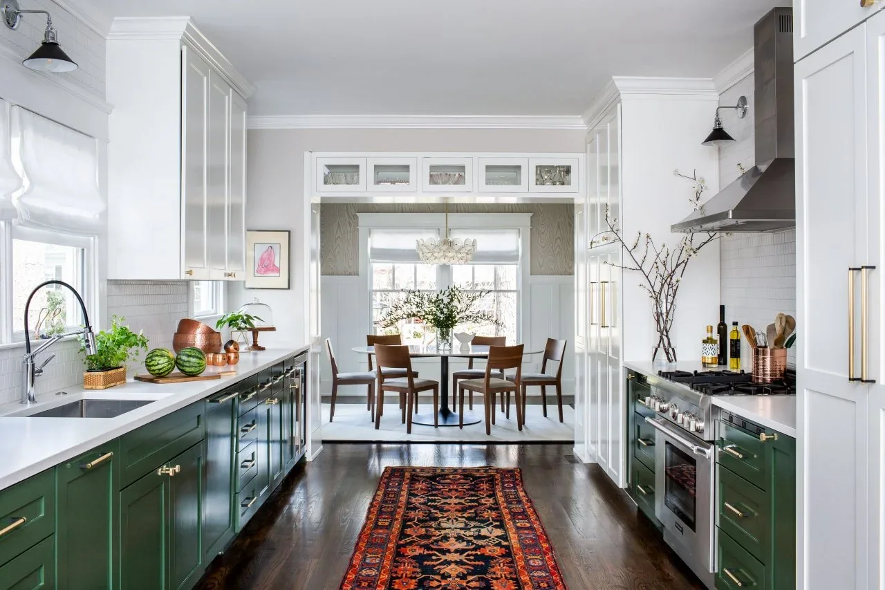 Deep green lower cabinets line a narrow kitchen beneath white upper and tall cupboards, with pale counters and a patterned runner.