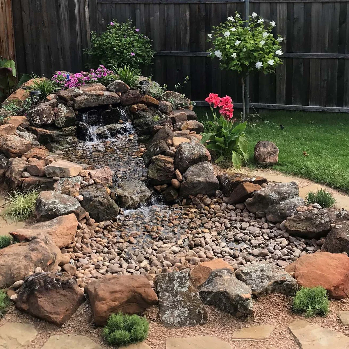 A low rock waterfall beside a small lawn, with a gravel receiving area, red and pink flowers and a dark wood fence.