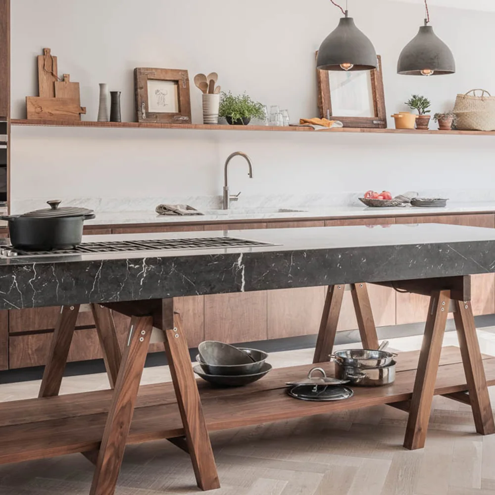 Walnut base cabinets behind a dark stone-topped trestle island contrast with an open lower shelf holding pots and lids.