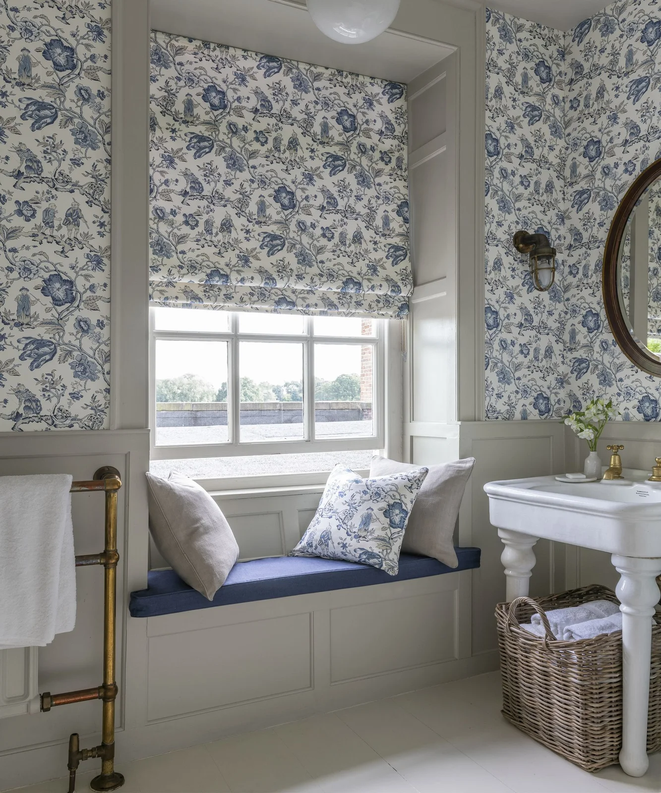 Blue-and-white botanical print covers bathroom walls, a window blind and cushion above a blue window seat beside a pedestal basin