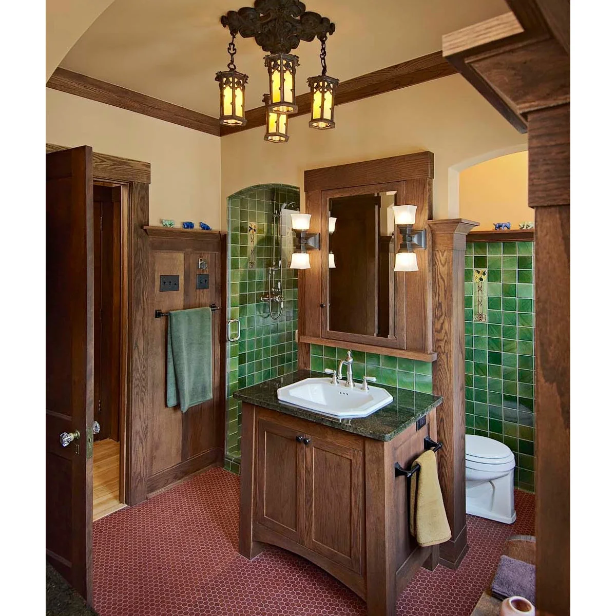 Green square tiles and decorative inserts surround a wood vanity and framed mirror above a small red-tile floor.