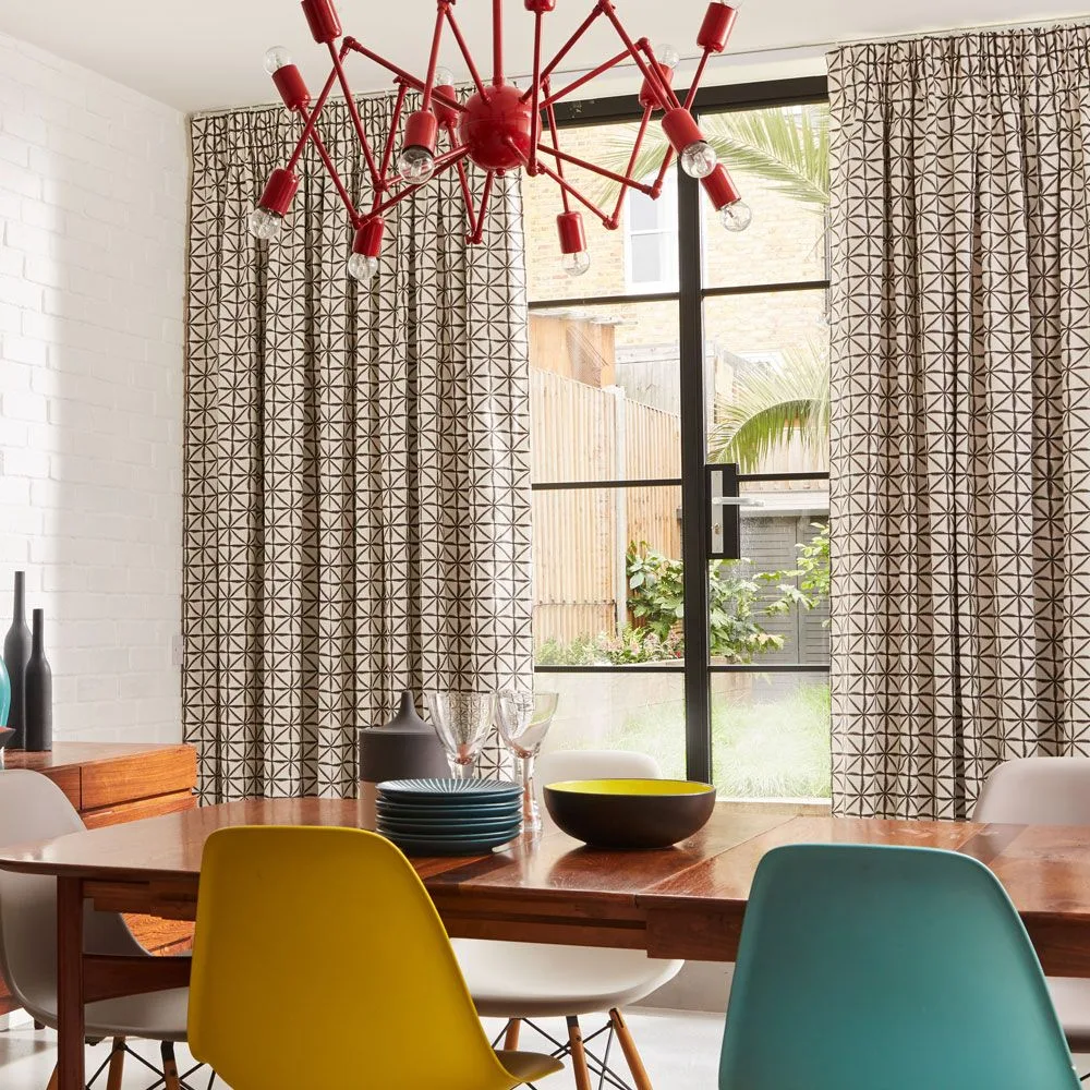 Black-and-pale geometric curtains around a black-framed glazed opening, with a wooden dining table, yellow and teal chairs, and a red angular ceiling light.