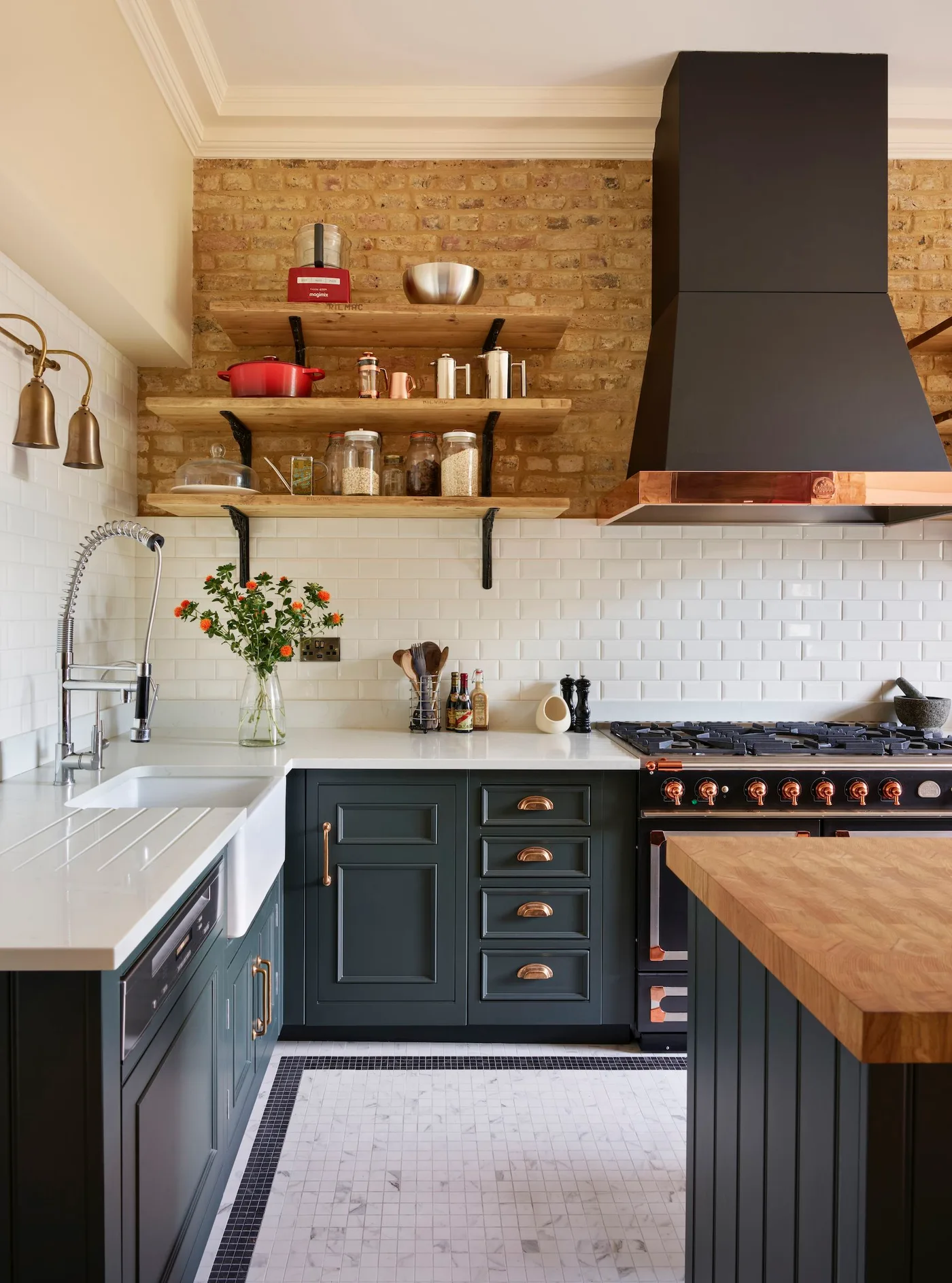 Kitchen corner with dark blue cabinets, white sink and tile, exposed brick, wooden shelves and tall black hood.