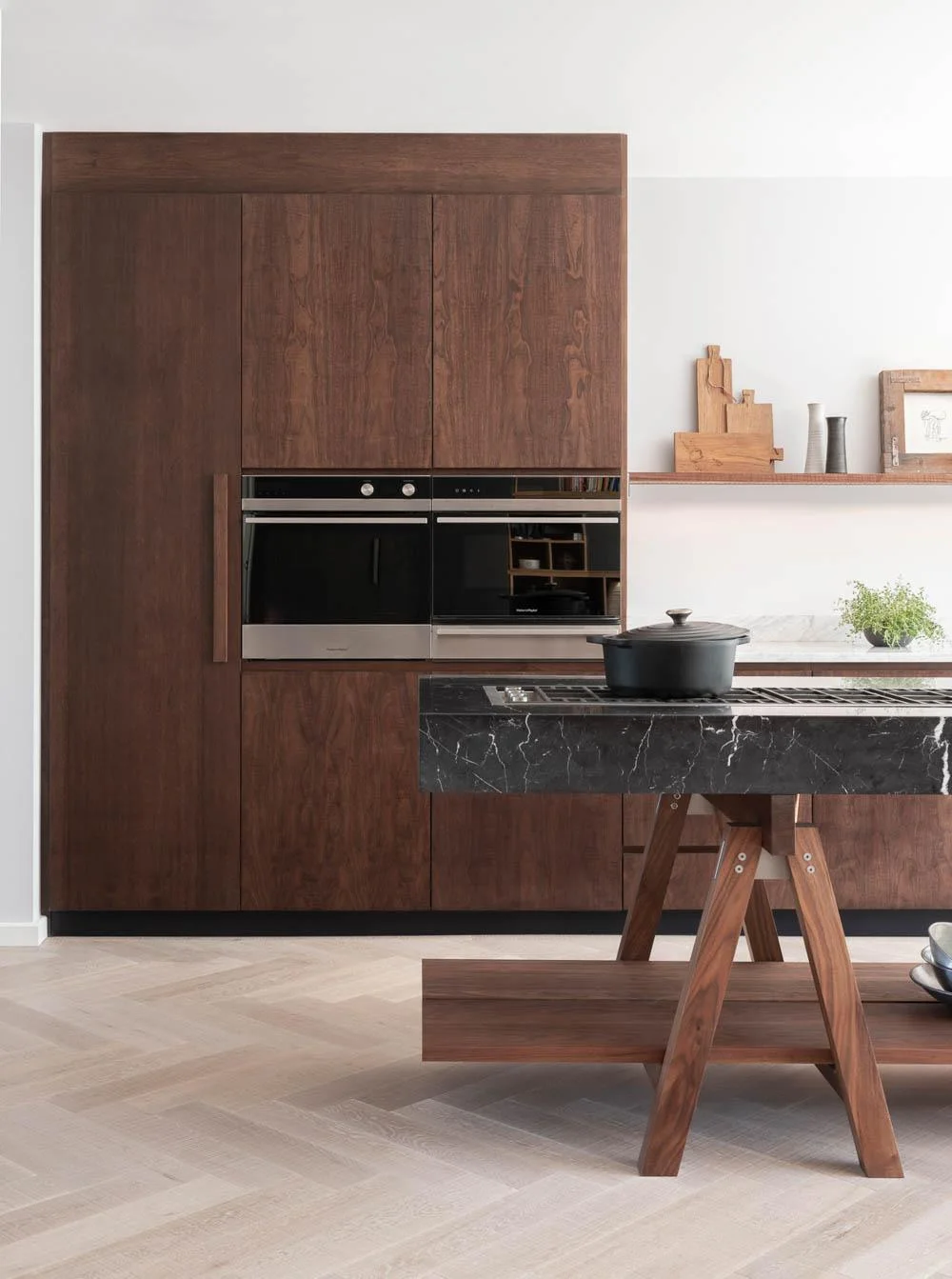 A flat-front walnut cabinet wall holds two black ovens, with pale walls and a dark veined island in front.