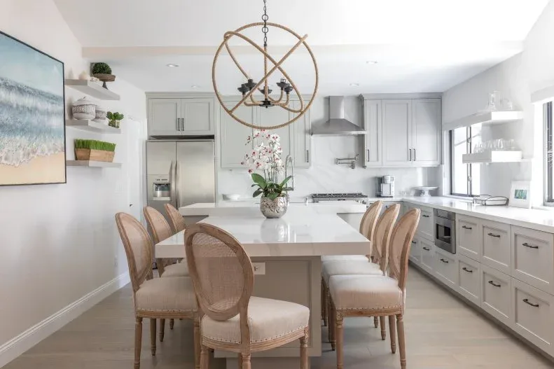 Pale eat-in kitchen with rectangular white table, six wooden woven-back chairs, coastal artwork and round open-frame chandelier.