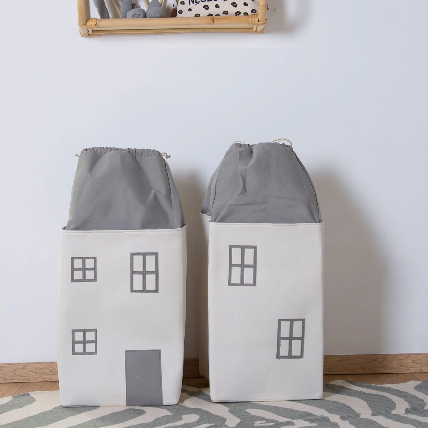 Two pale house-shaped fabric storage containers with gray drawstring tops on a wood floor