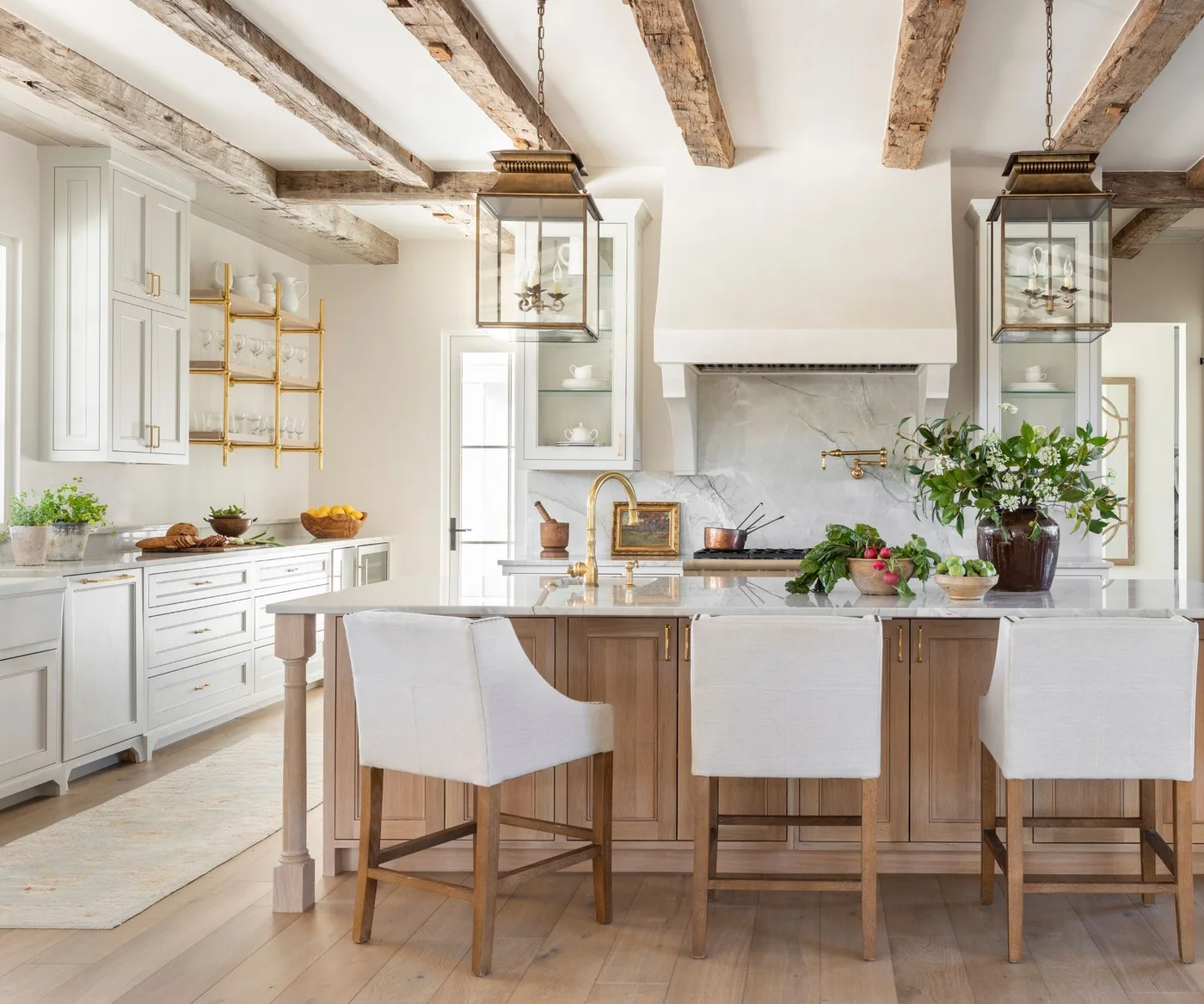 A white framed kitchen has exposed ceiling timbers, a wood island, large glass lanterns, upholstered stools and a broad pale cooker hood.