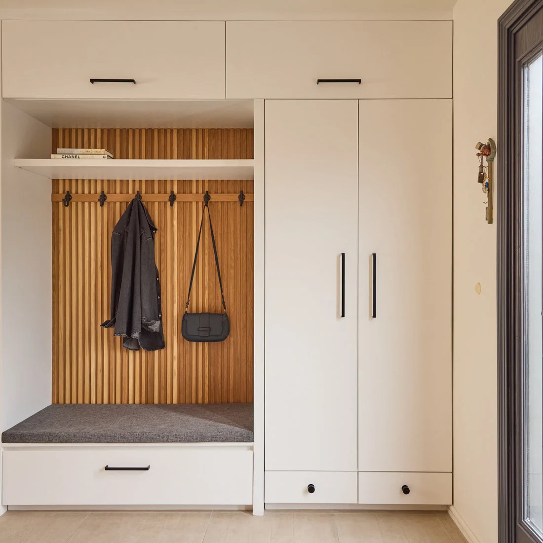 A gray-cushioned bench and wood slat hook panel sit beside tall white cupboards with black handles and a lower drawer.