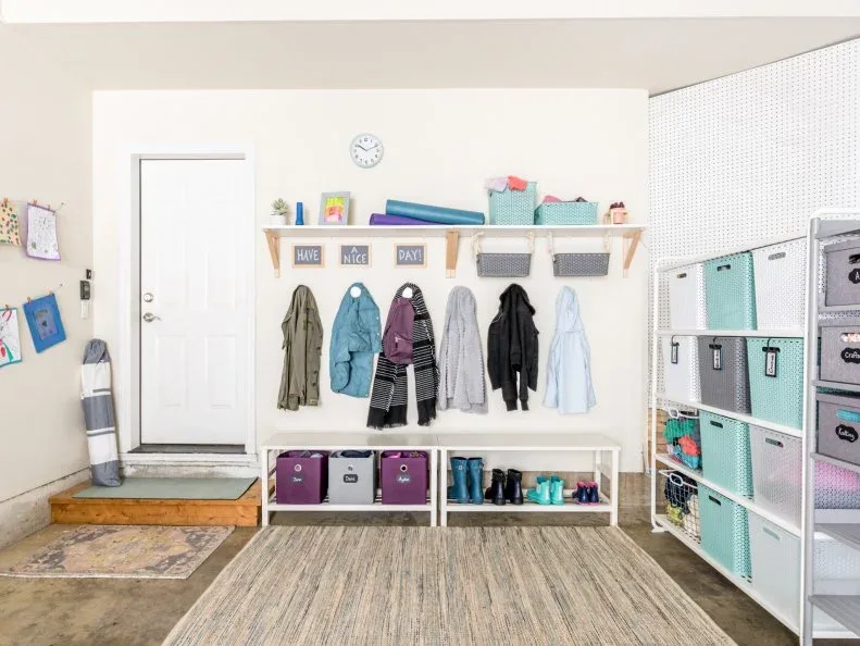 Garage entry wall with coats above white shoe cubbies, a high shelf and colored storage bins beside a door.