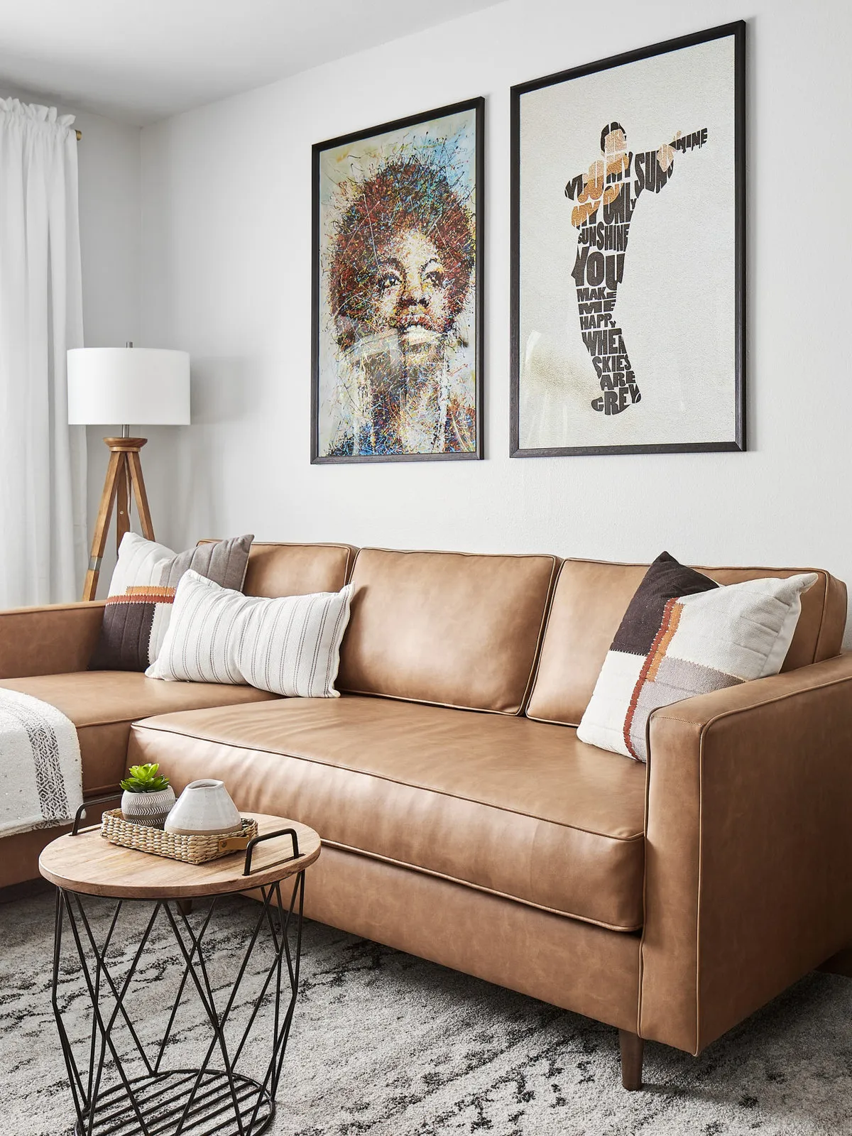 Tan leather sofa beneath two large black-framed pictures beside a wooden tripod lamp and wire-based side table.