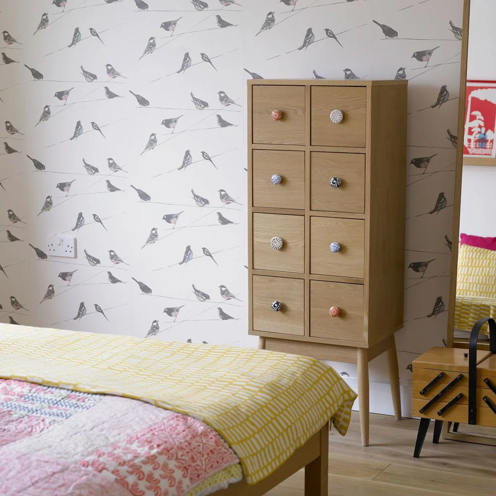 Gray birds on fine lines repeat across white wallpaper behind a tall wood drawer cabinet with colorful knobs, beside yellow and pink bedding.