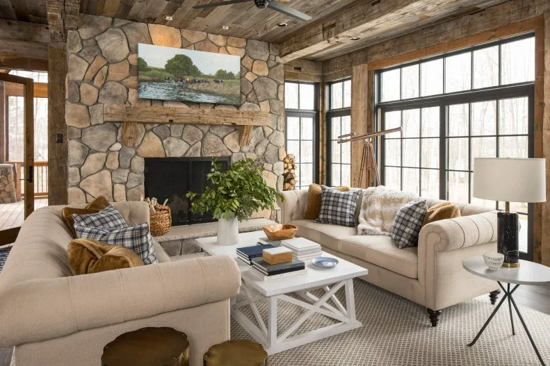 Irregular pale and brown stones frame a fireplace beneath a weathered timber mantel in a beamed living room.