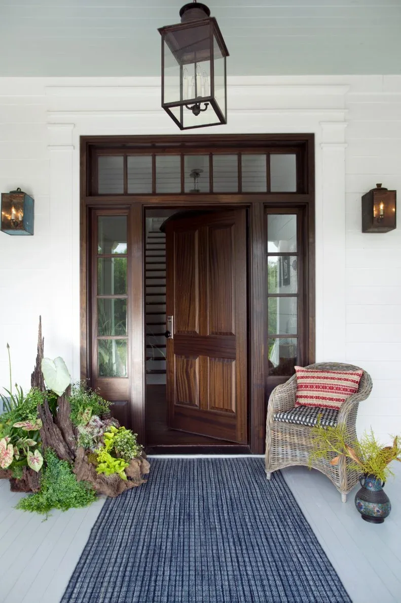 Wood country door with glazed sidelights, pale porch boards, hanging lantern, woven chair and planted arrangement