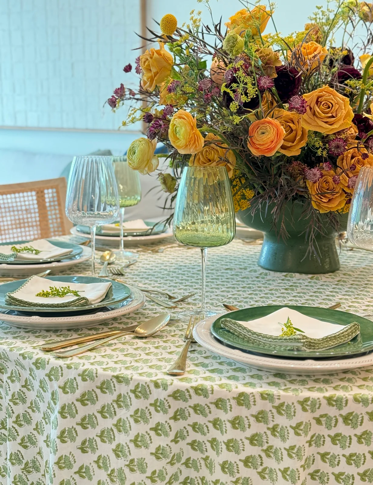 Green patterned cloth and green plates sit beneath orange flowers with pale napkins and clear glasses