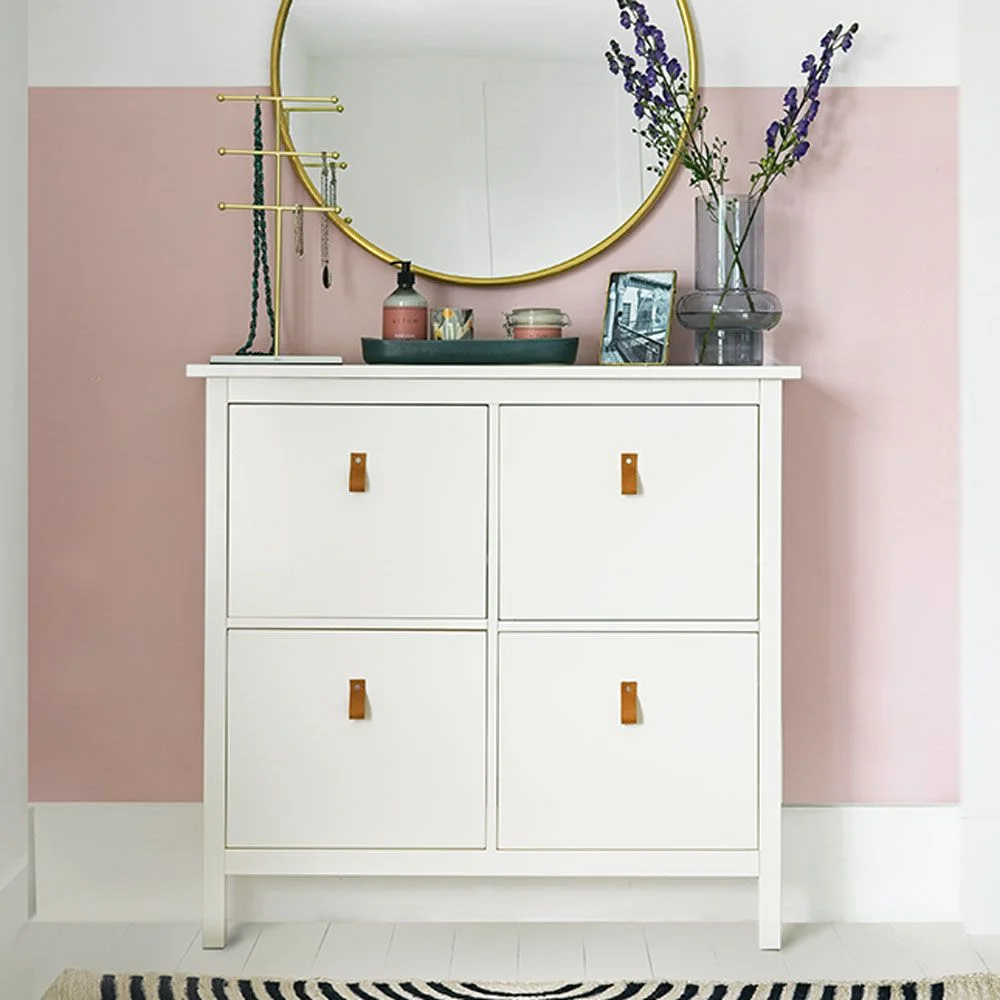 White shallow shoe cabinet with tan tab handles below a round mirror on a pink wall