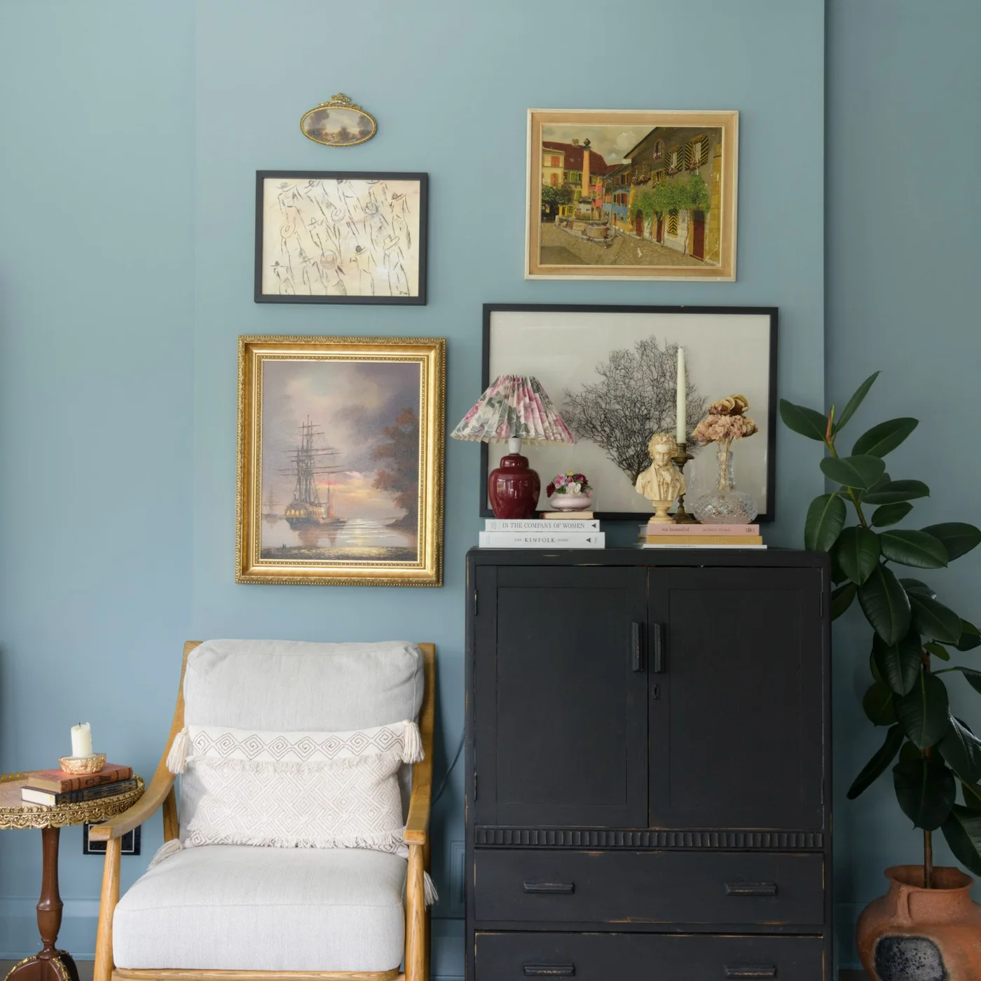 Mixed framed pictures hang above a dark cabinet beside a cream timber-framed chair on a pale blue wall.