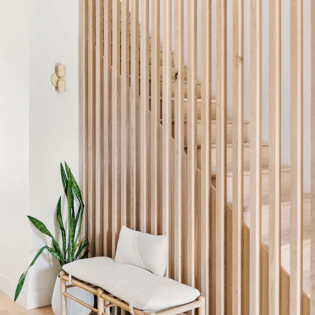A pale cushioned wood bench stands beside a tall wood slat screen with a stair visible behind it and a small wall light nearby.