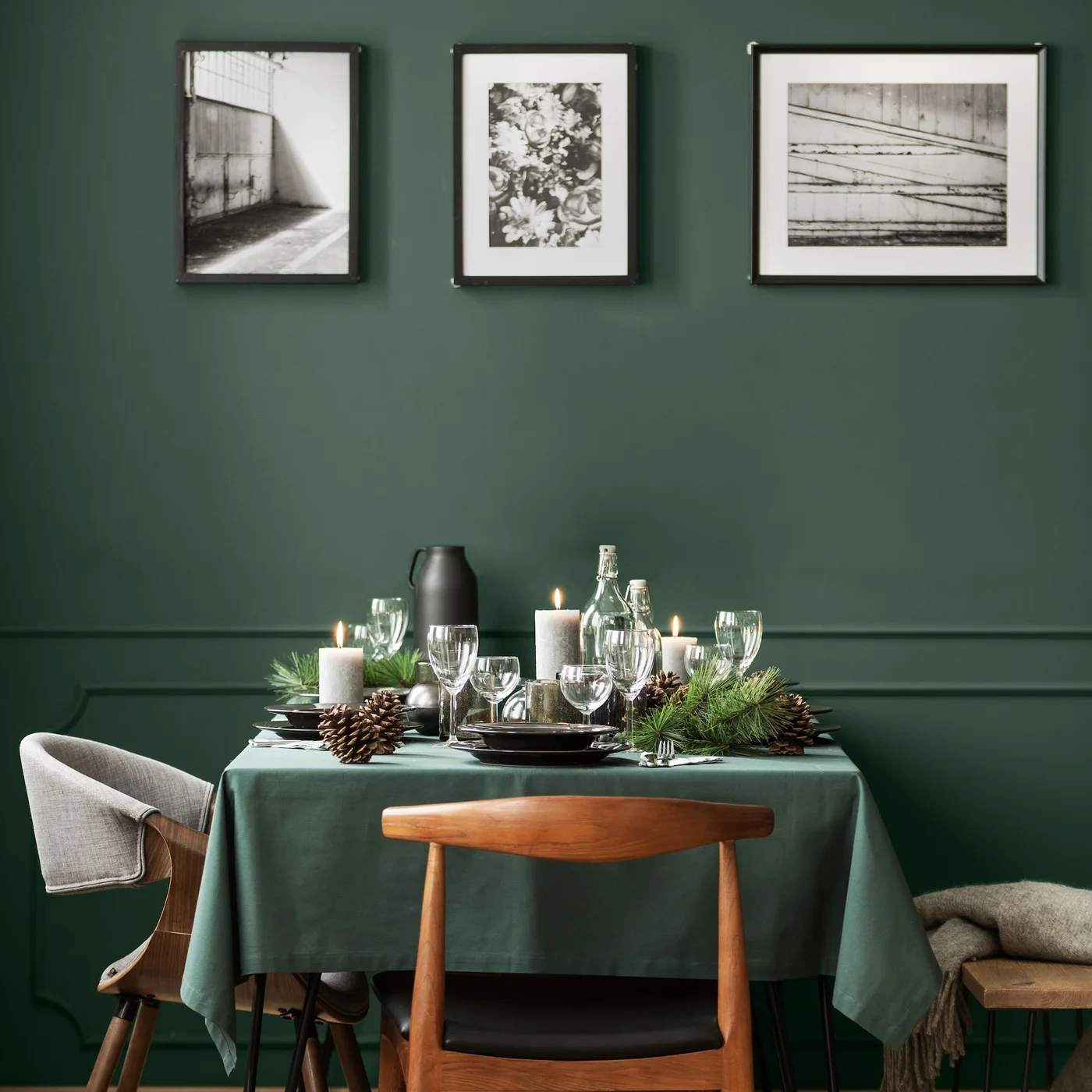 A green-clothed table stands against a deep green wall beneath three framed monochrome pictures with mixed chairs, glassware, candles and pine greenery