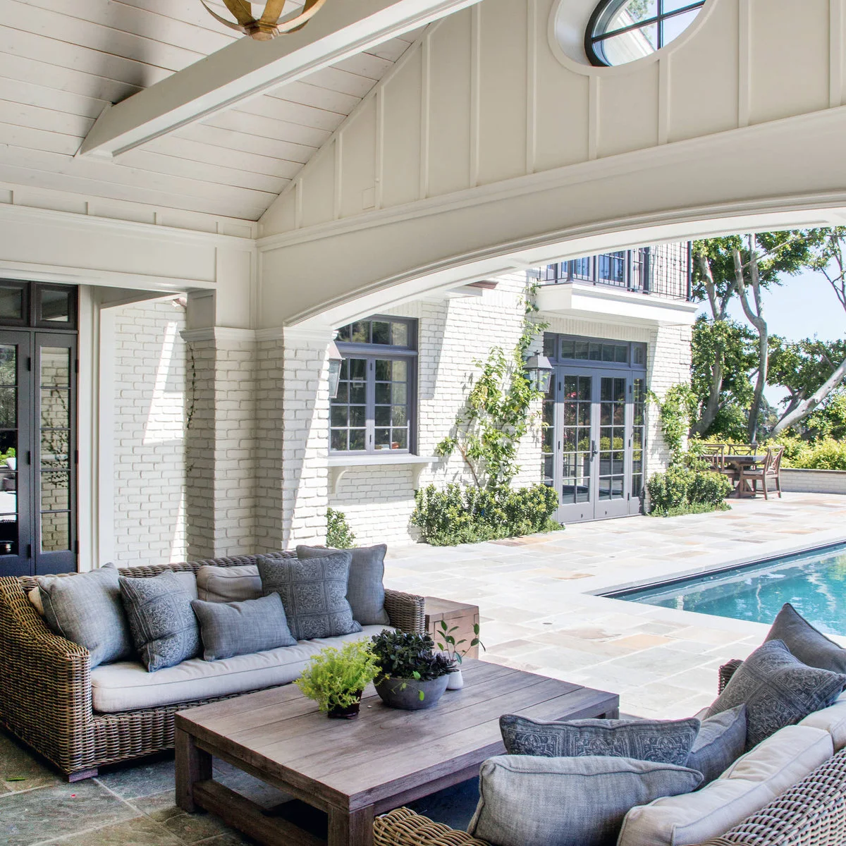 A pale arched beam frames gray-cushioned woven seating and a broad wood table beside a pool.
