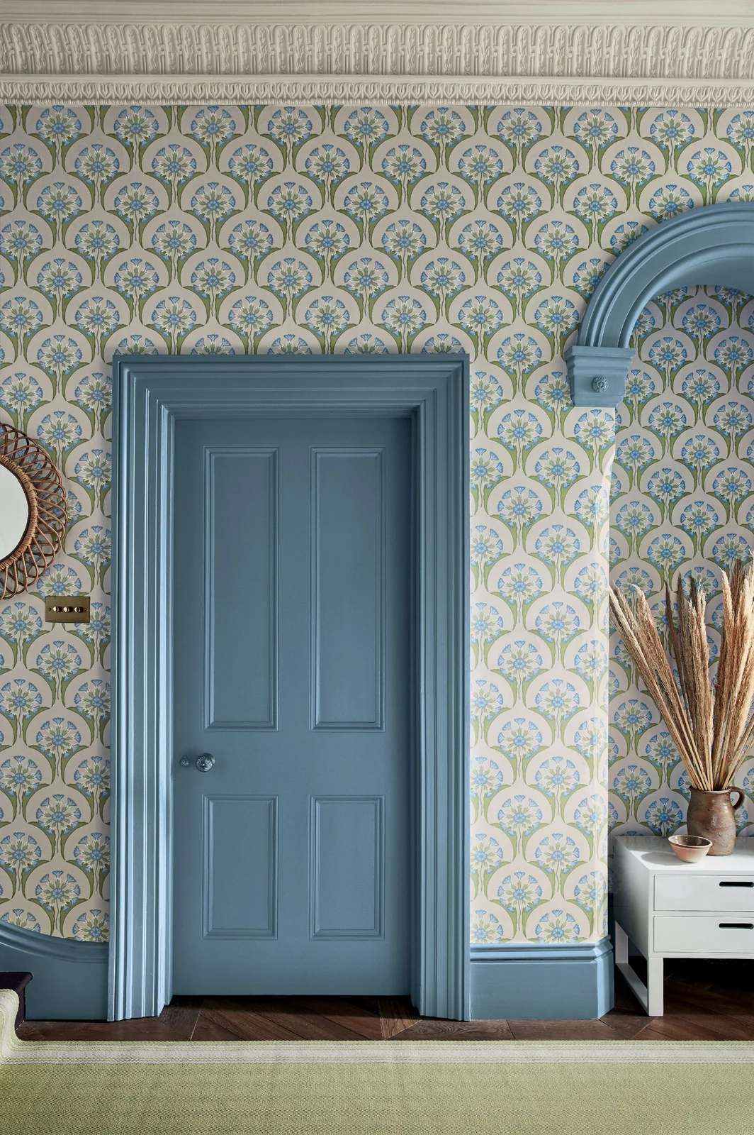 Blue painted door trim and an arch sit against a repeated blue-and-green floral wall beside dried stems and a green runner