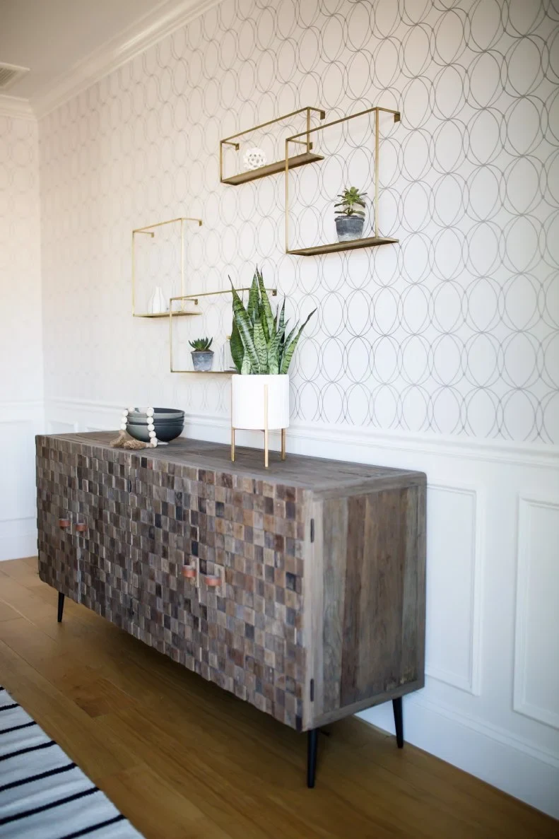 A dark block-textured sideboard holds a black bowl and a raised white plant pot beneath gold-colored open box shelves on patterned wallpaper.