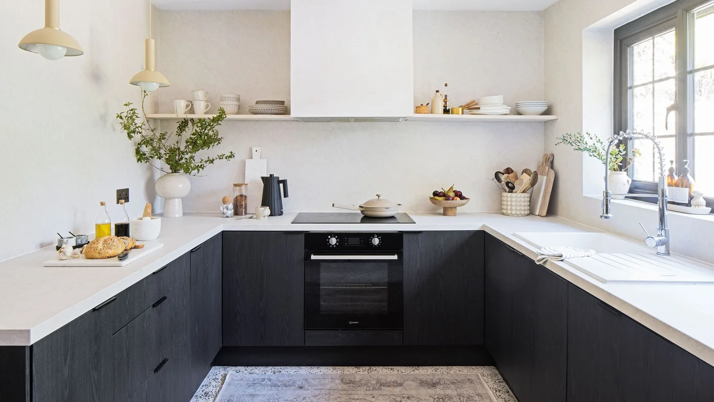 A U-shaped kitchen has dark plain lower fronts, pale counters and walls, a long shallow shelf, two small yellow pendants and a window above the sink.