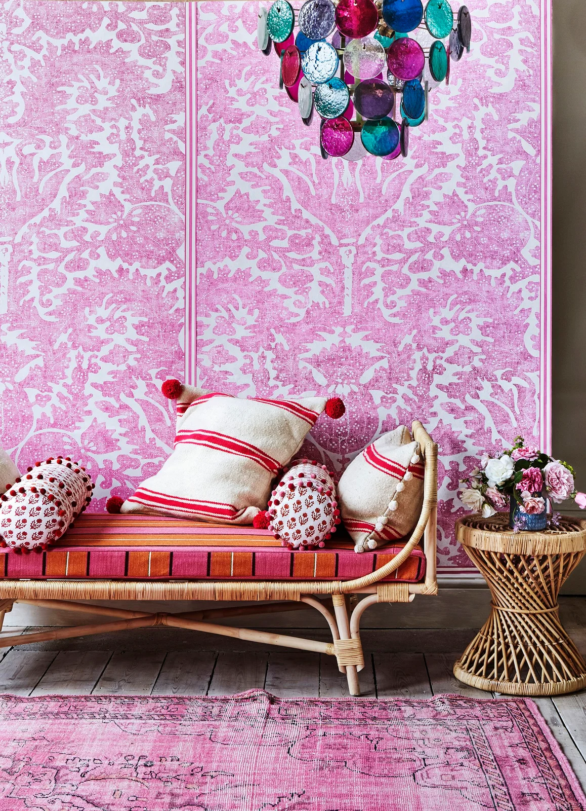 Pink patterned wallpaper panels stand behind a woven sofa with striped cushions and a multicolored pendant.