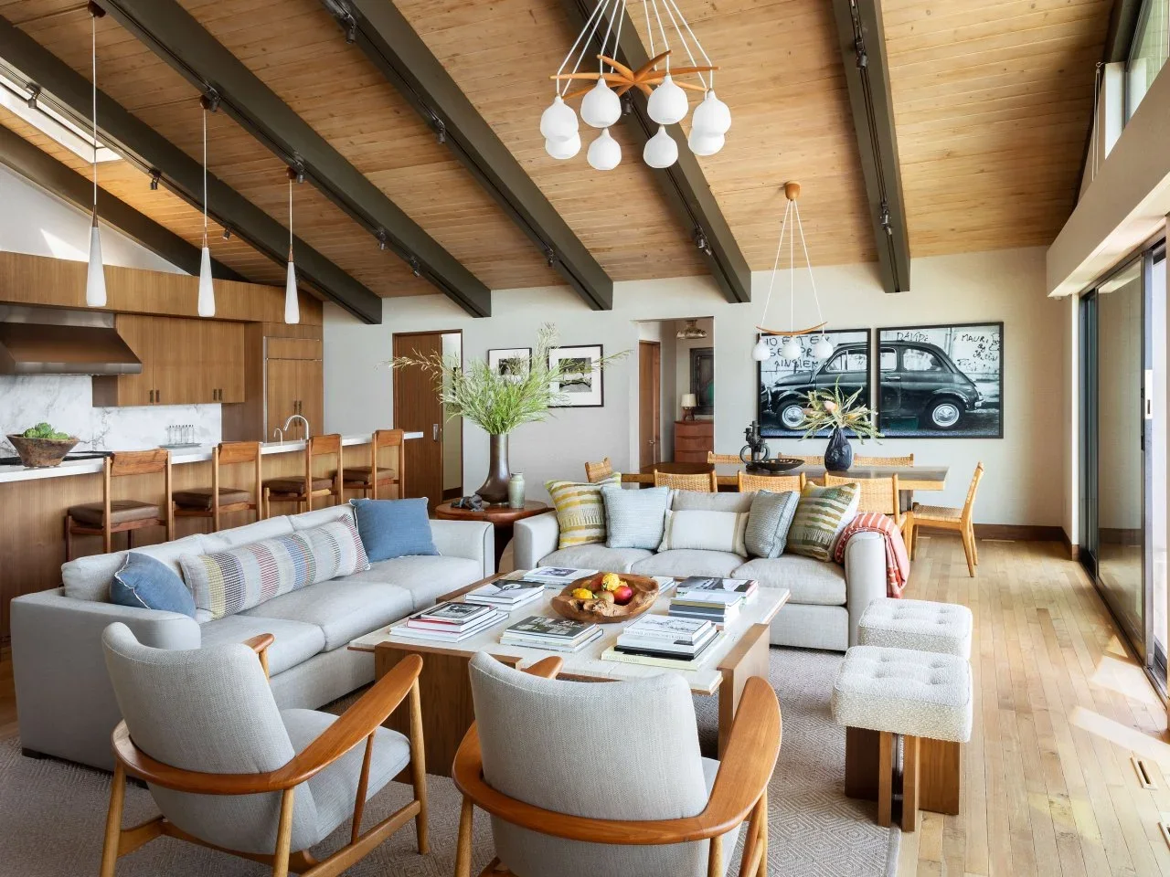 Pale sofas and timber chairs form a seating group beside an open timber kitchen and dining table under exposed beams.