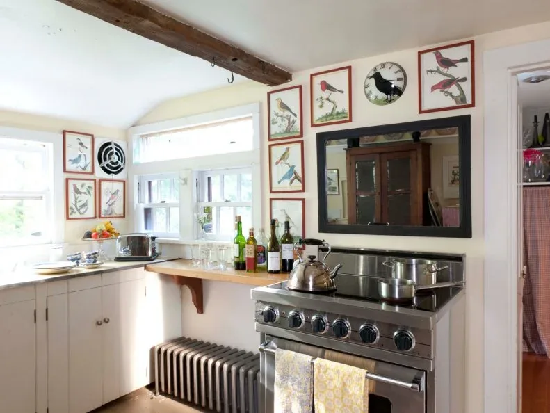 Pale kitchen with framed bird pictures around windows and above a dark-backed range area beneath an exposed beam