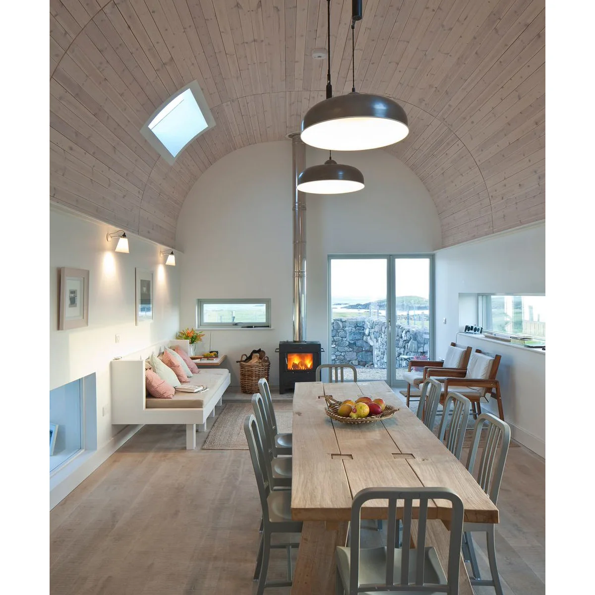 Narrow room beneath a curved pale wood ceiling with a long dining table, fitted bench, armchairs and a stove beyond.