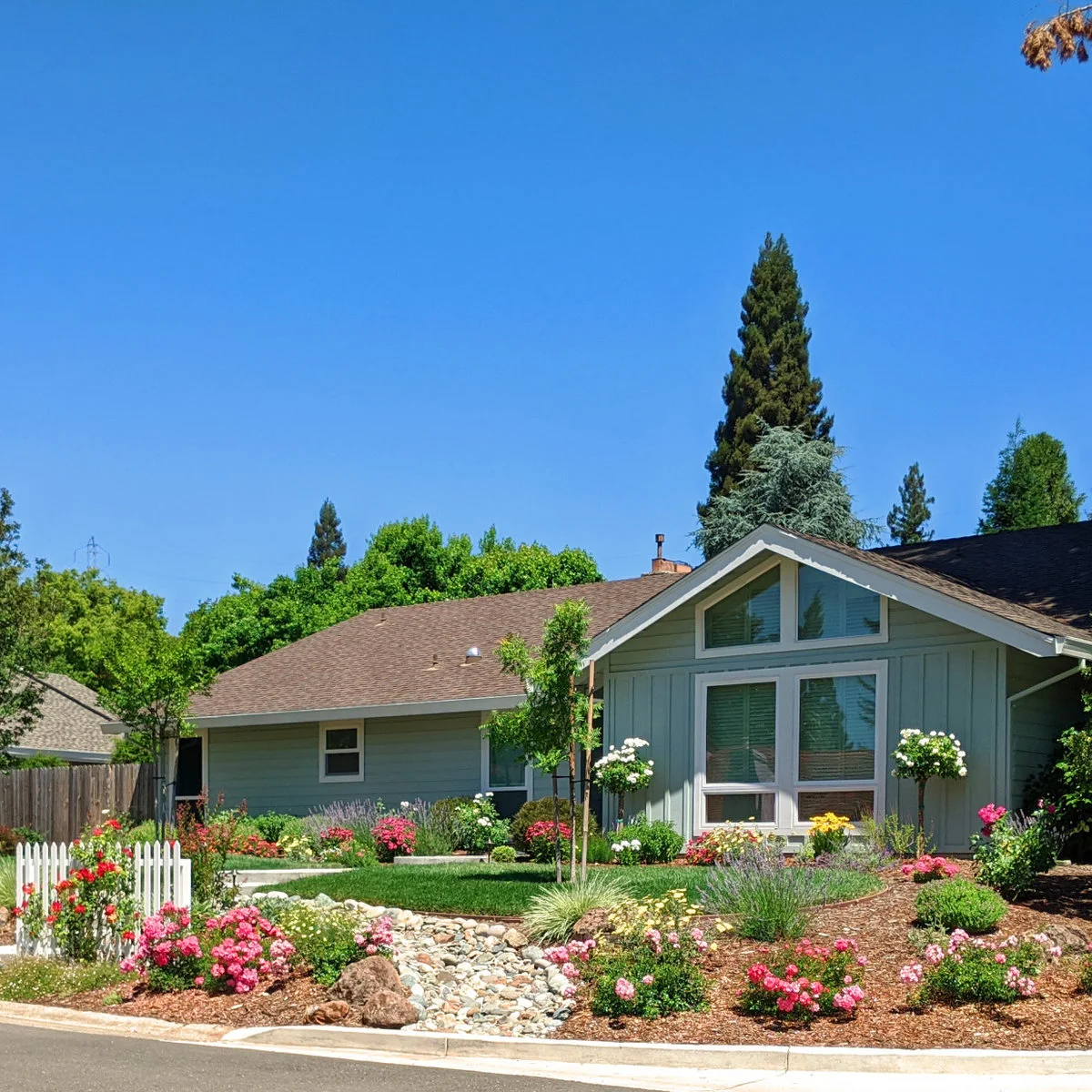 A blue-gray bungalow has a compact lawn, partial white picket fence and curved flower beds with a band of rounded stones.