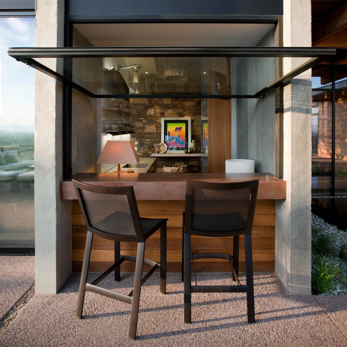 Raised glass serving window above a brown counter with two dark outdoor stools and wood cladding below