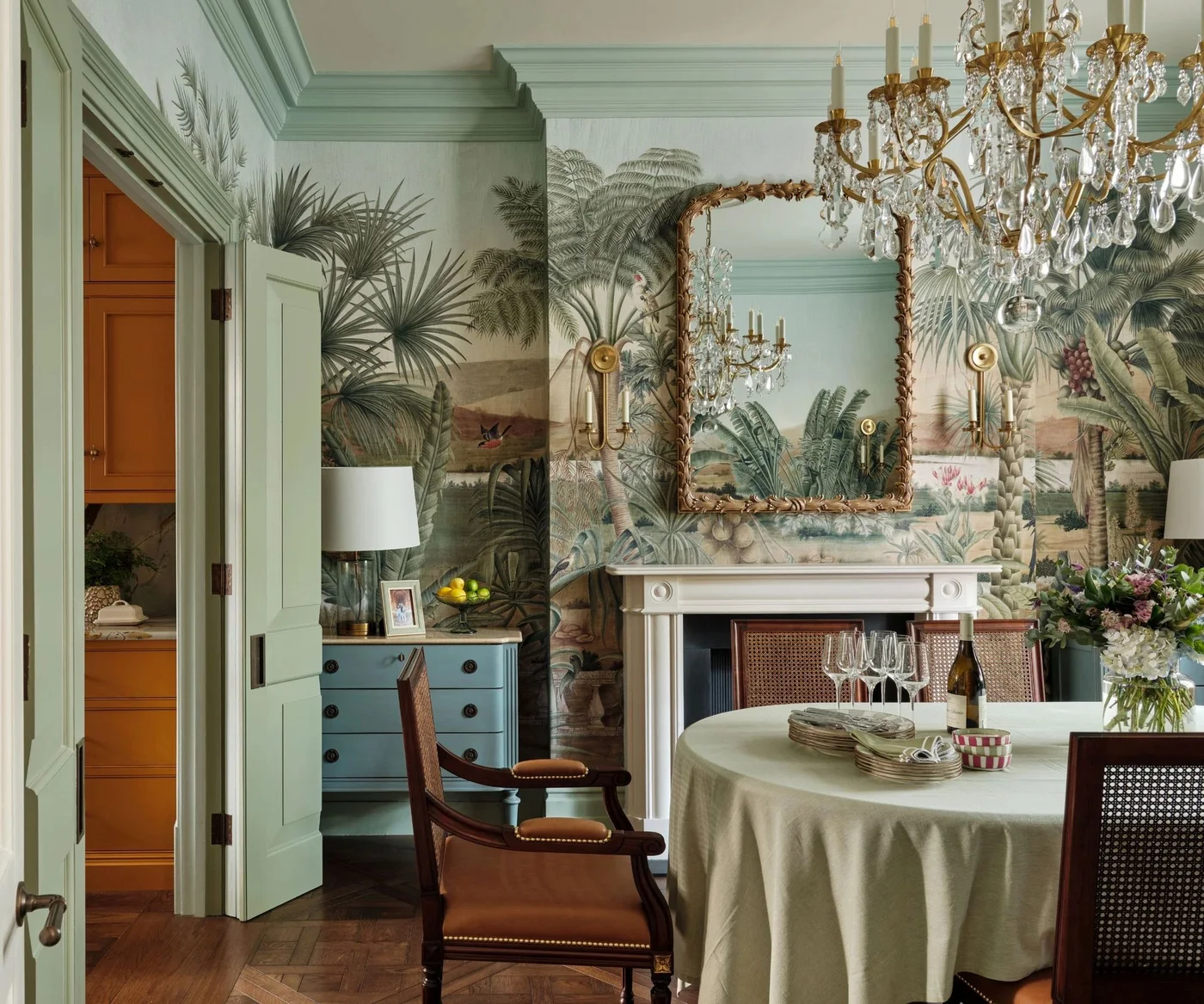 A tropical scenic mural surrounds an ornate mirror and chandelier above a round cloth-covered table beside pale green trim and dark woven-back chairs