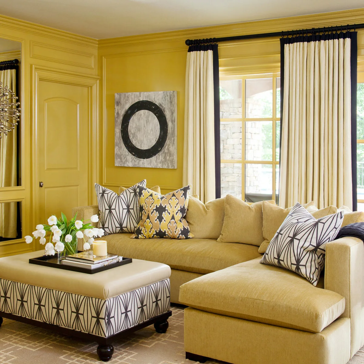 Living room with yellow walls and sectional sofa, black-and-white patterned cushions and pale curtains with dark borders