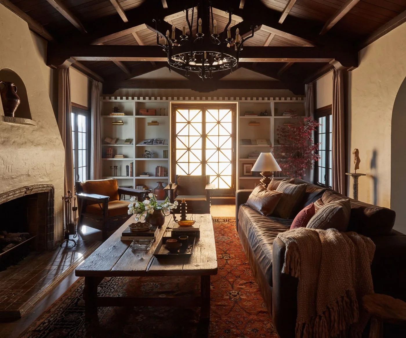 Dark living-room seating and a long wooden table sit beneath exposed wood beams beside pale open shelves, a patterned red rug and textured fireplace