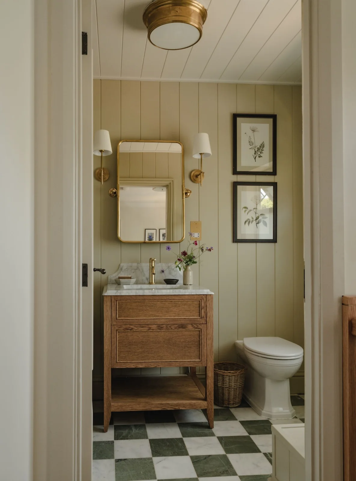 Pale olive paneling frames a wood vanity, gold-edged mirror and botanical pictures above an olive-and-white checked floor.