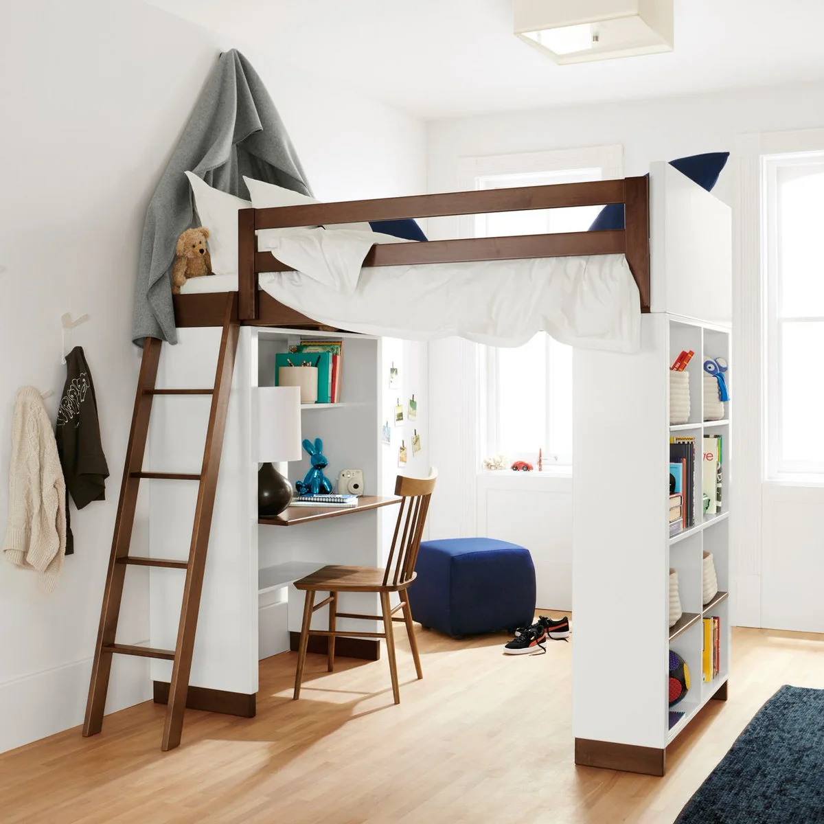 White loft bed with brown rails and ladder, bookcase ends, a desk below and a blue ottoman near the windows.