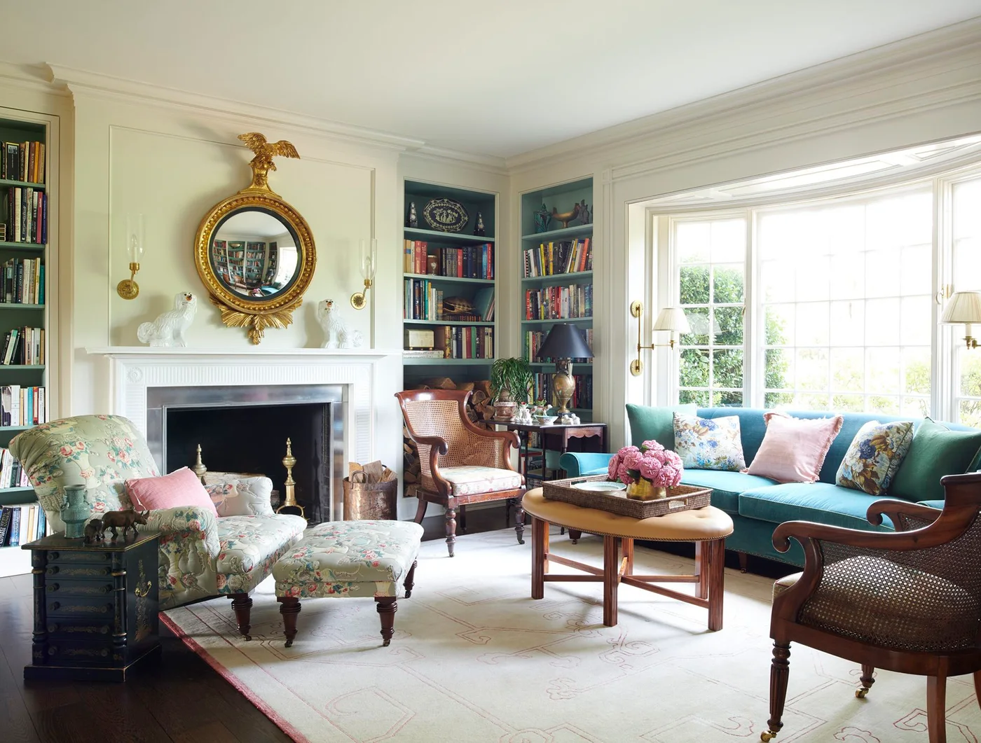 A white fireplace stands between dark-backed bookshelves beneath a round gold mirror in a room with patterned chairs and a green sofa.