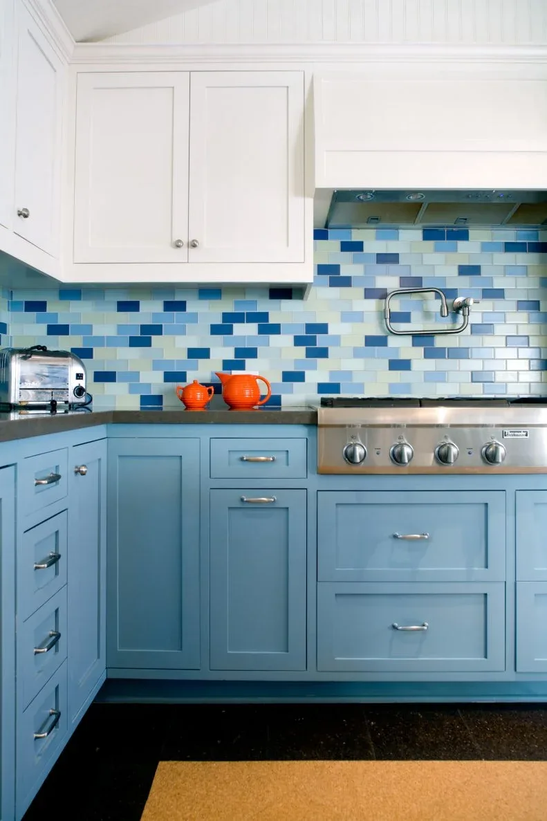 Kitchen with pale blue lower cabinets, white upper cabinets and varied blue, cream and white rectangular backsplash tiles behind the cooker.