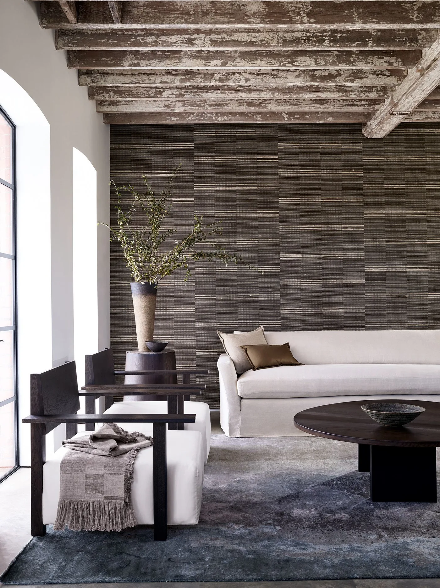 Dark horizontally patterned wall beneath a textured wood ceiling with pale sofa, dark wood armchairs and round table