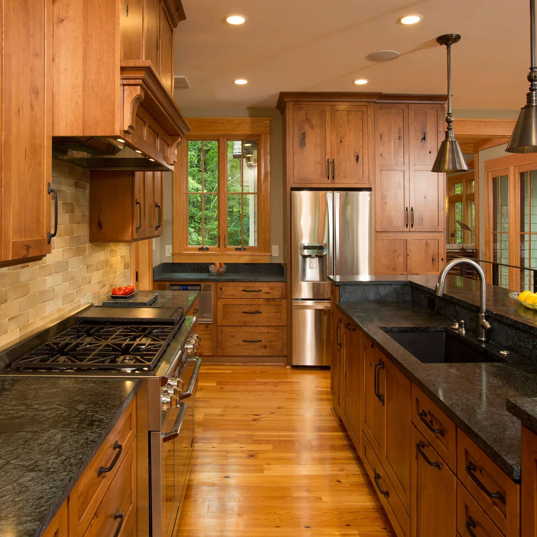 Wood cabinets and flooring around a dark topped island beside a pale tiled cooking wall