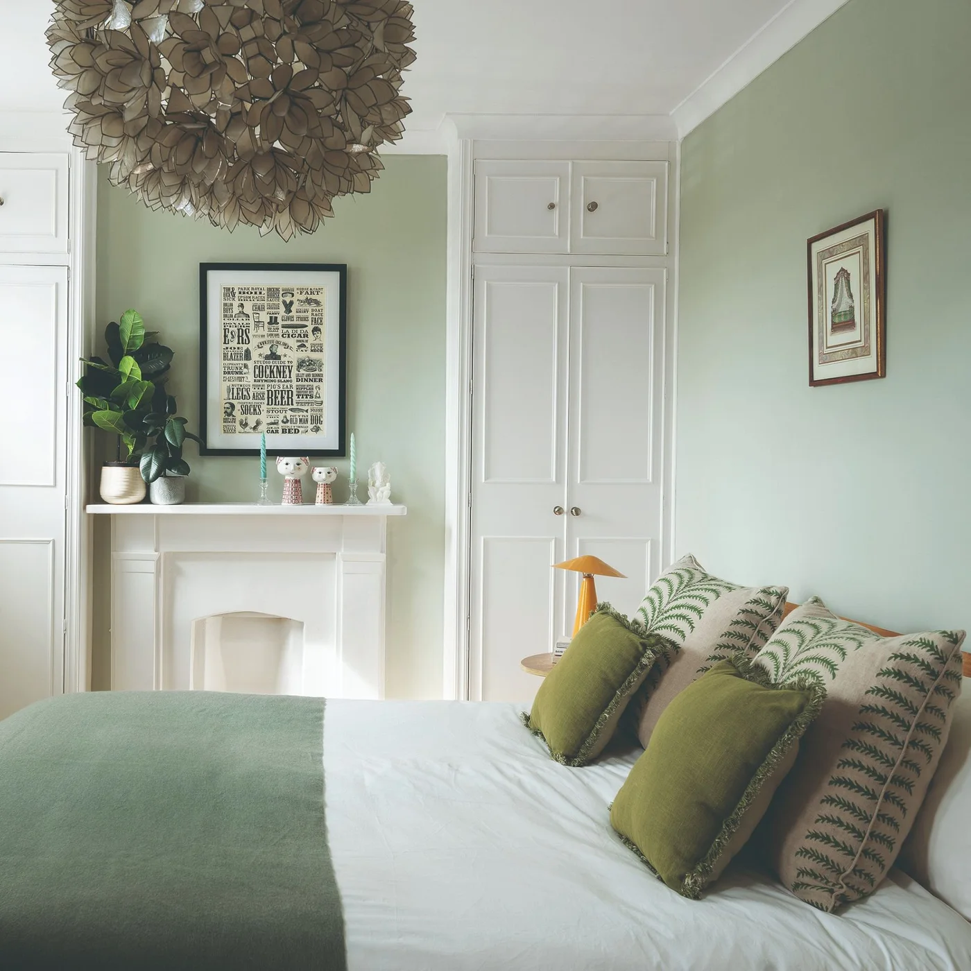 Light sage bedroom walls sit between white fitted cupboards and a white fireplace beside green cushions and bedding.