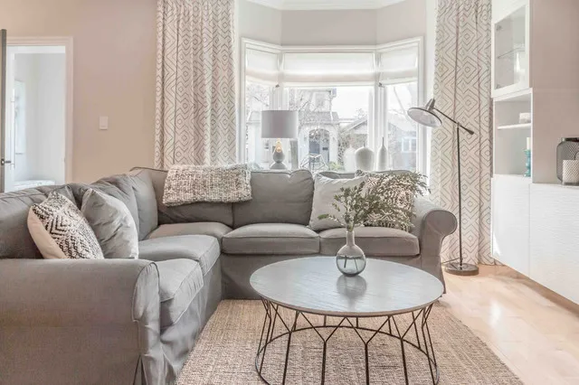 A light gray corner sofa sits beside a broad bay window and a round table with an open dark frame.
