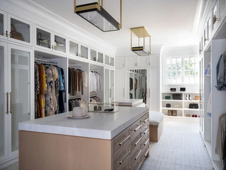 A white walk-in closet has glass-front upper cupboards, open hanging bays and a timber drawer island.
