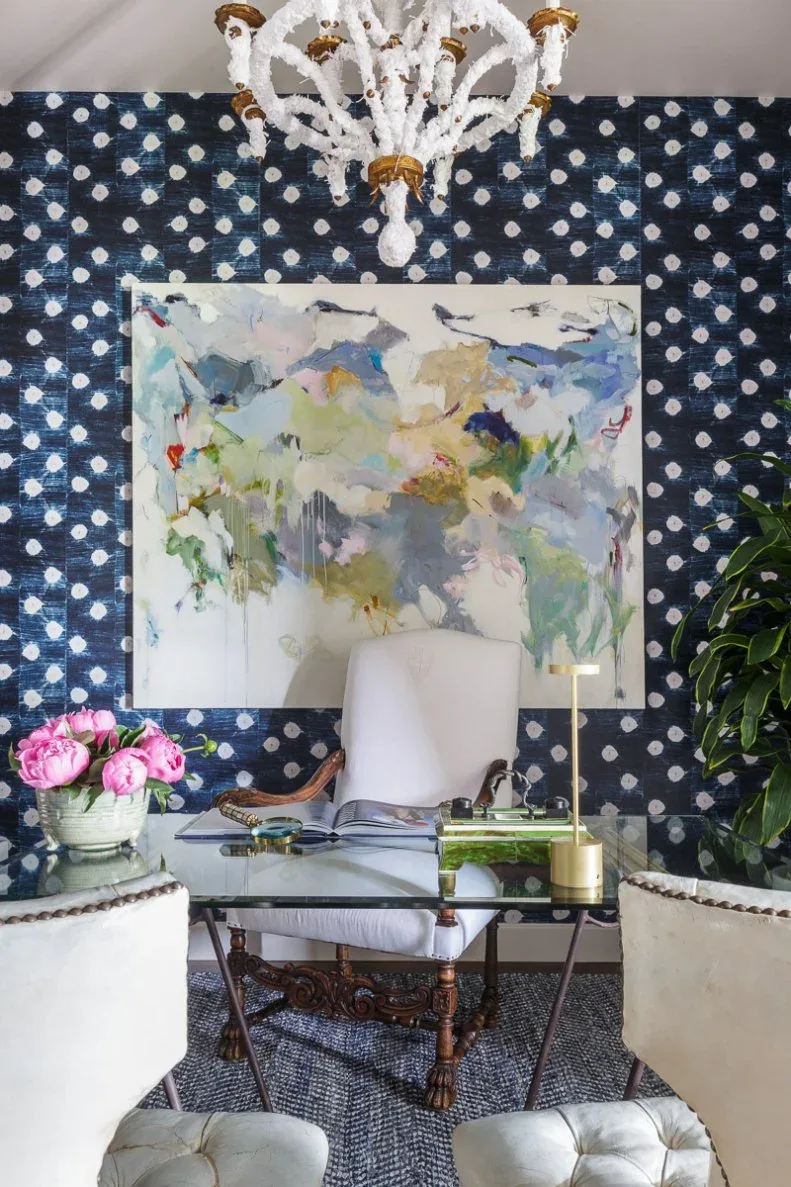 Home office with dark blue dotted wallpaper, large colorful abstract artwork, clear-topped ornate wooden desk, pale chair, patterned rug and leafy plant.