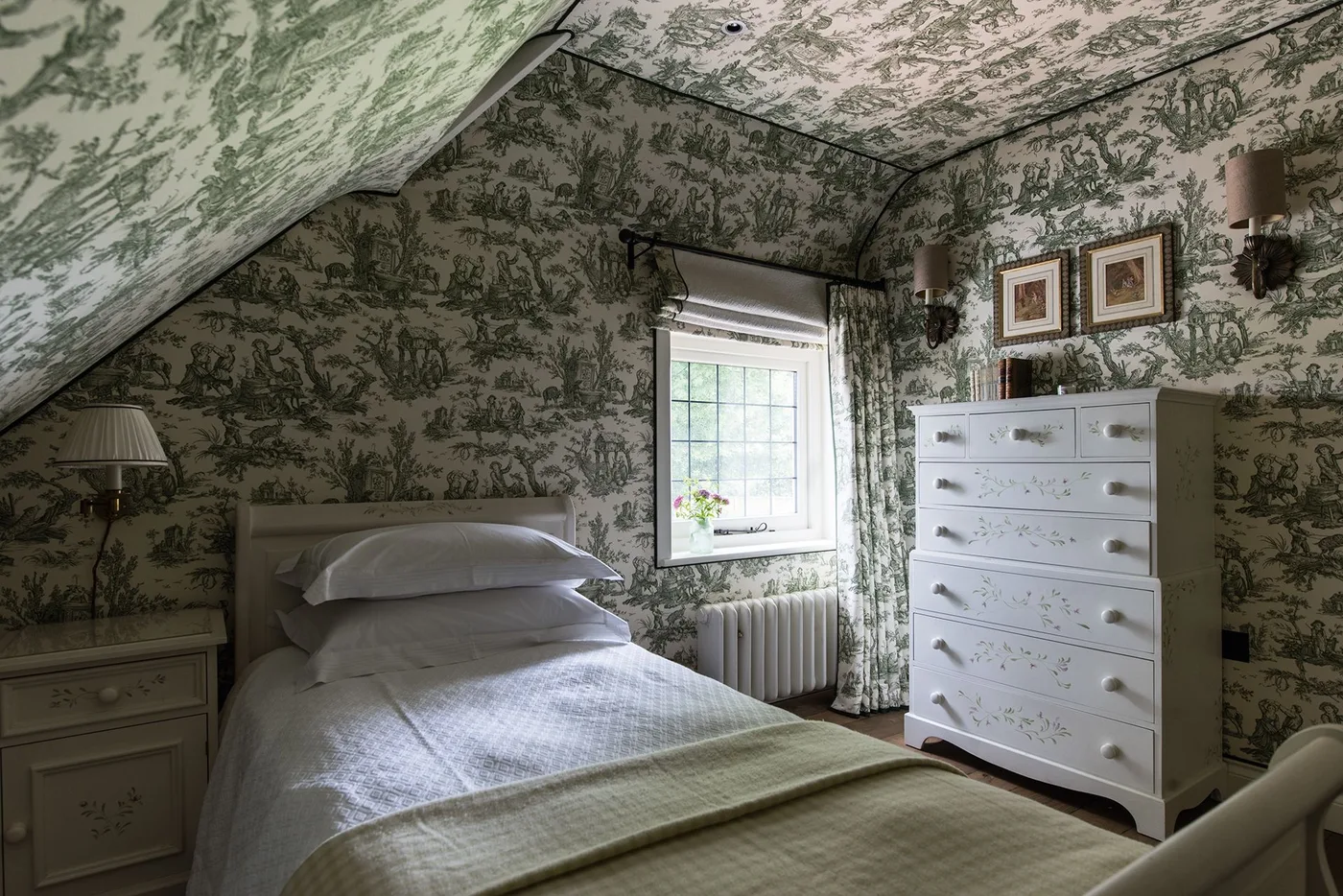 Sloping-ceiling bedroom with green scenic wallpaper across walls and ceiling, a pale bed and tall white chest of drawers.