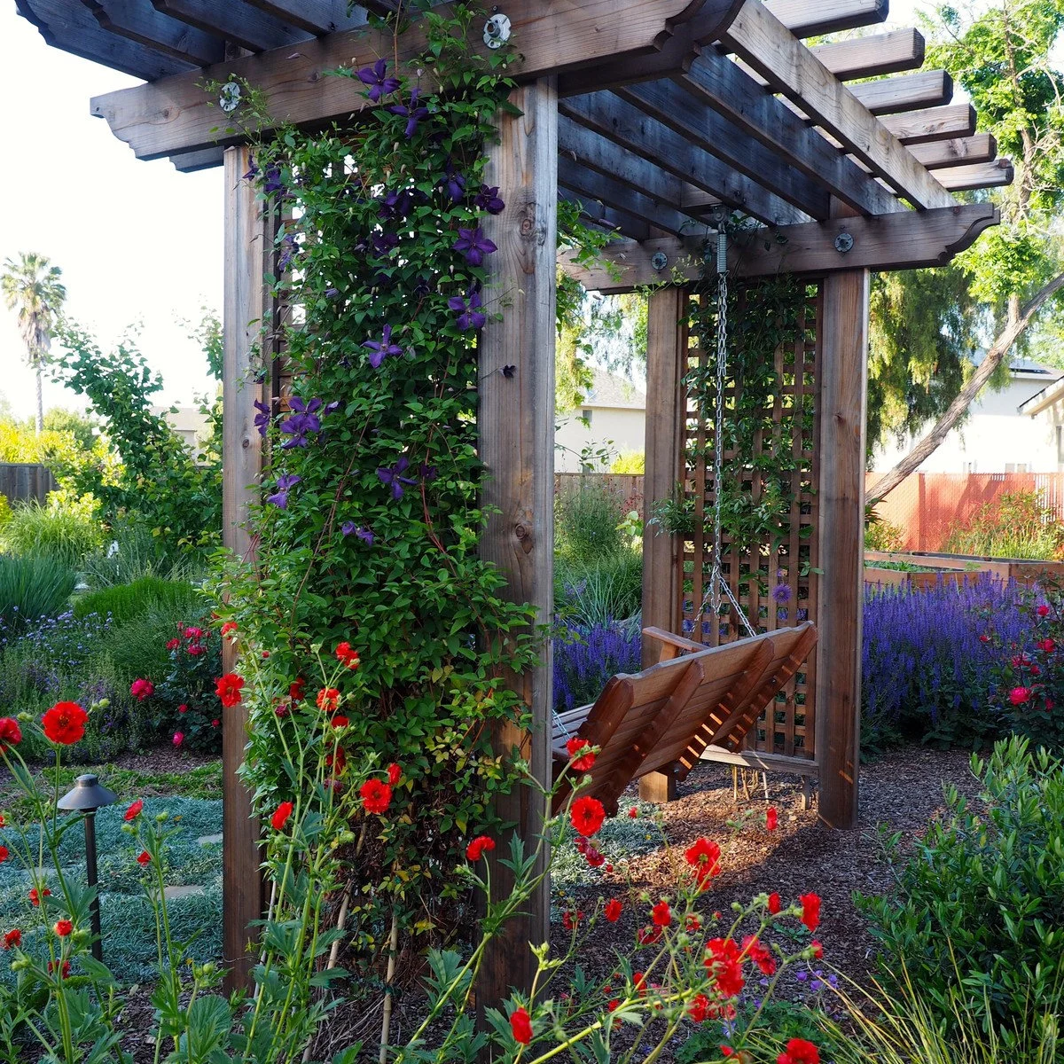 A wood swing hangs within a substantial timber arbor surrounded by climbing plants and red and purple flowers.