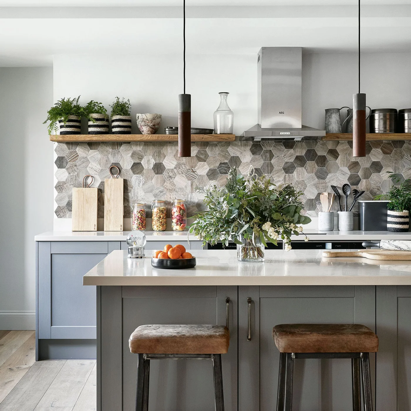 Mixed gray hexagonal tiles cover a kitchen backsplash beneath a wooden shelf beside gray cabinets and a pale island